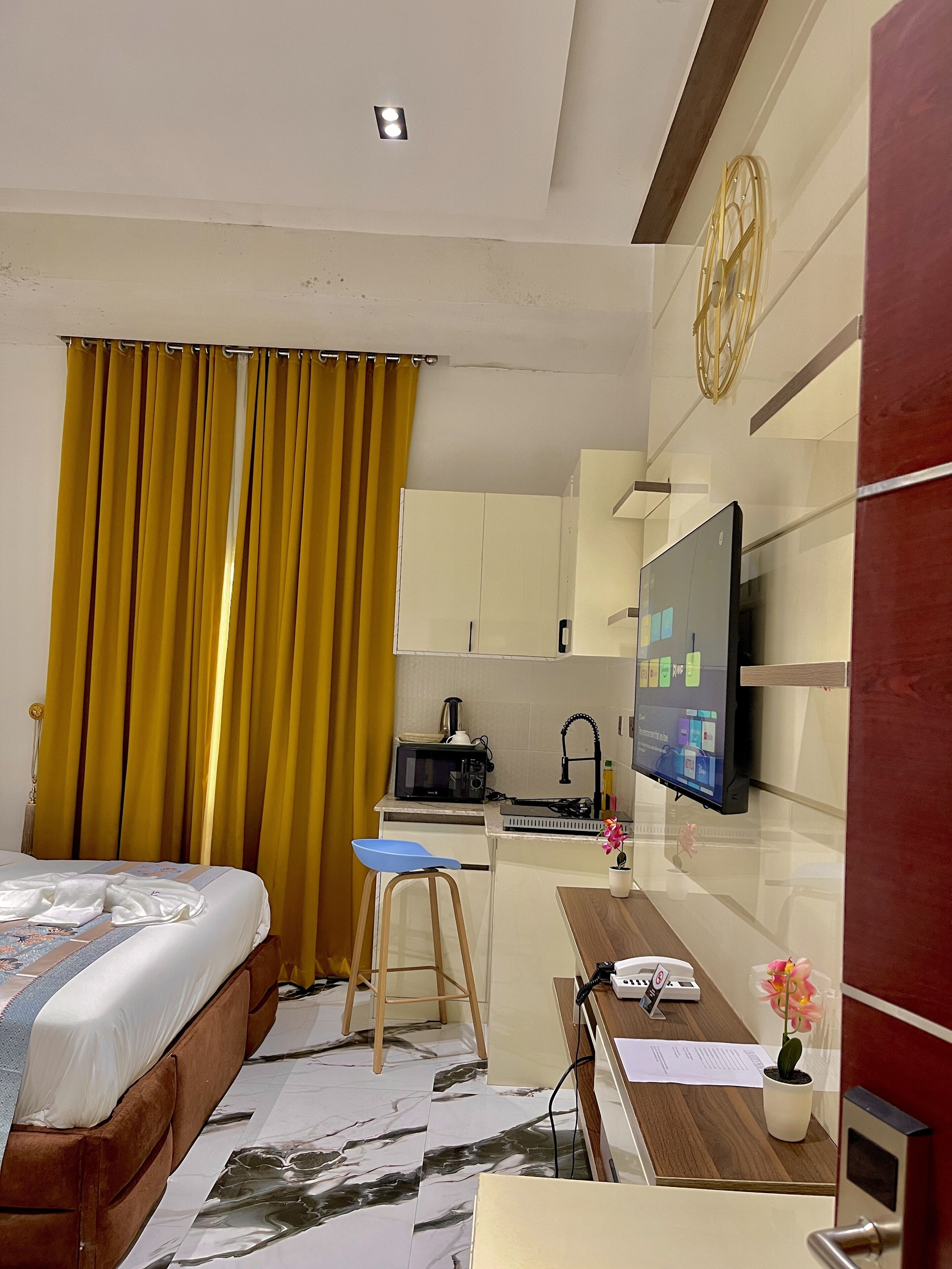 A hotel room with a bed, a desk with a computer, a TV, a small kitchen area, and a bathroom.