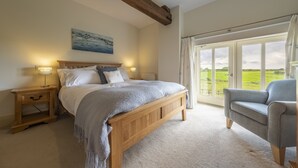 A bedroom with a wooden bed, bedside tables, a view of the countryside through large windows, and a grey armchair. - 6 Courtyard Barn, Docking, Norfolk (Docking)