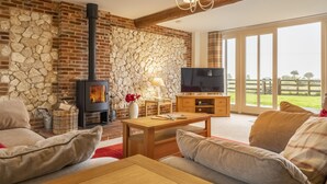 A cozy living room with a stone wall, a fireplace, a television, and a large window with a view of the outdoors. - 6 Courtyard Barn, Docking, Norfolk (Docking)