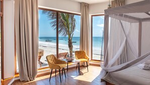 Un dormitorio con un ventanal grande que da a una playa, una cama con dosel y dos sillas mirando a la ventana.
