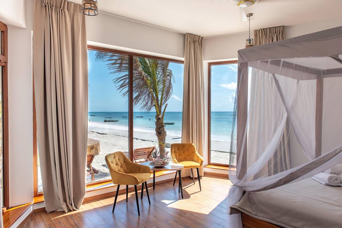 Un dormitorio con un ventanal grande que da a una playa, una cama con dosel y dos sillas mirando a la ventana.