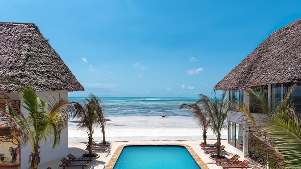 Un área de piscina con sillones de descanso y palmeras, que da a una playa con vista despejada del océano.