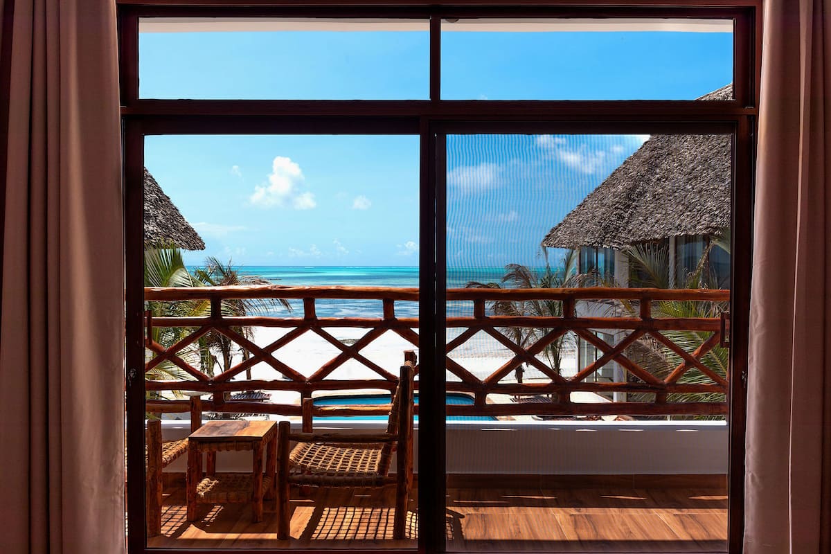 Un balcón con piso de madera, una mesa y una silla, con vistas a una playa con chozas de techo de paja y un cielo azul despejado.