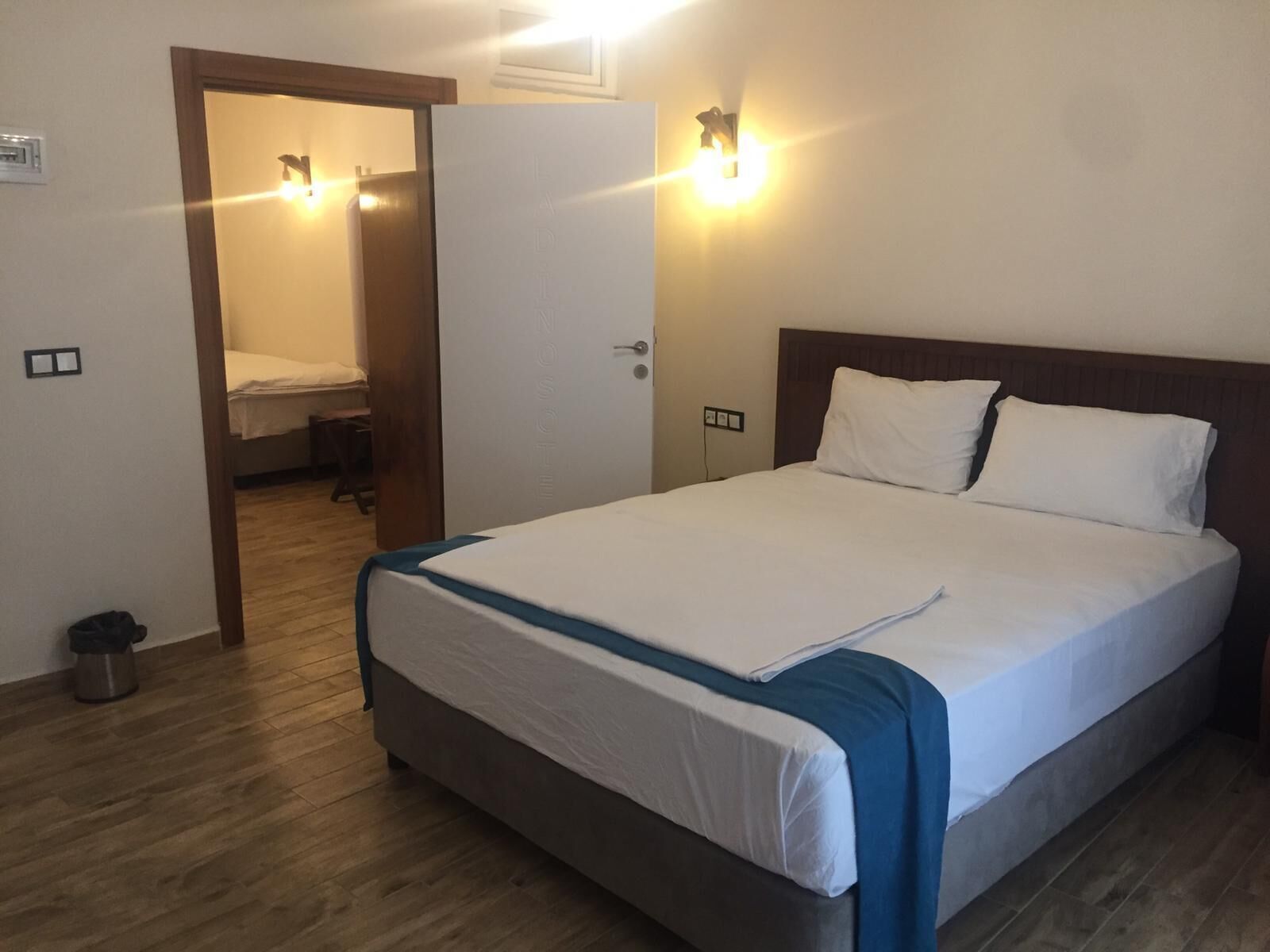 A hotel room with a bed, a wooden floor, a door, and a wall-mounted light fixture.