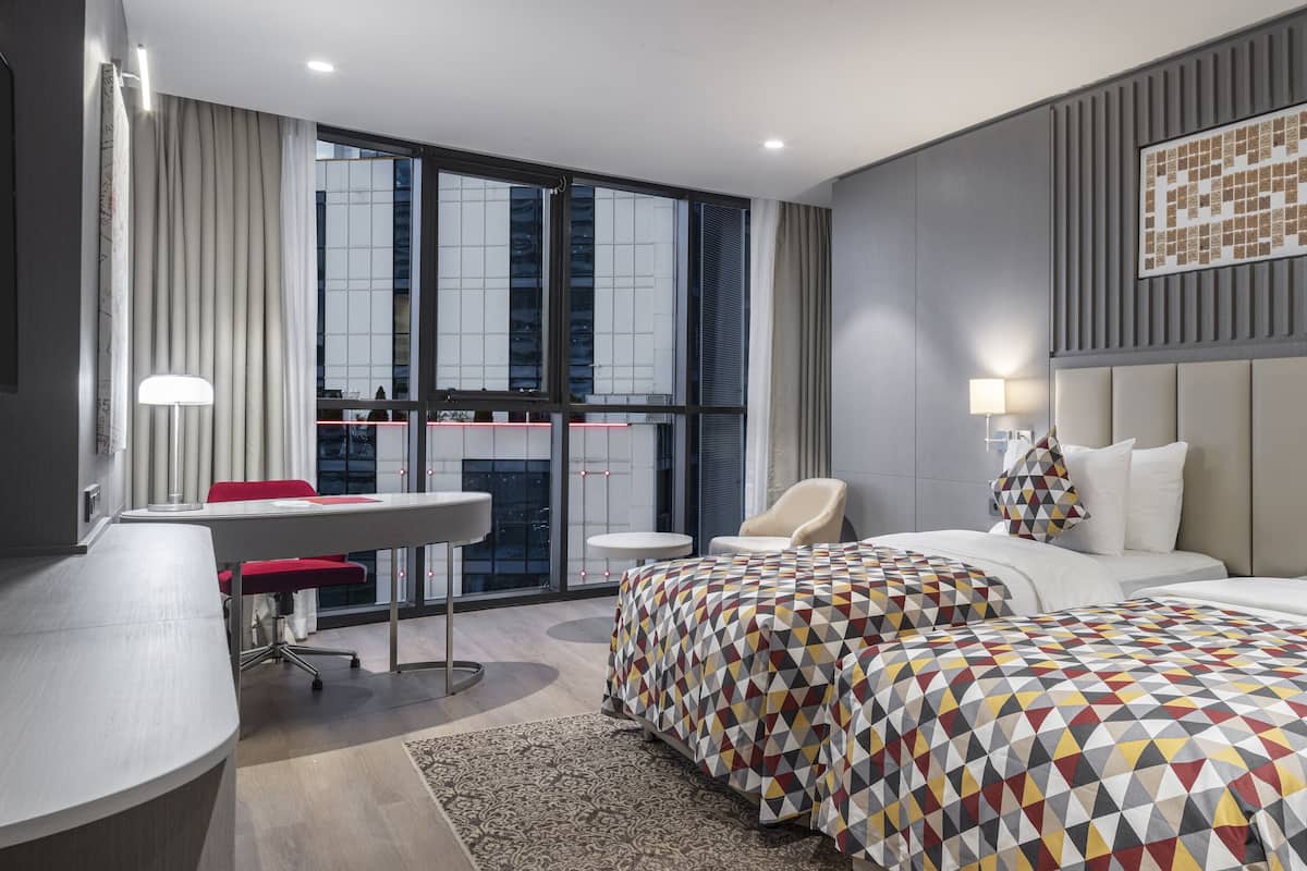 Une chambre d’hôtel moderne, dotée d’un grand lit, d’un bureau et offrant une vue sur la ville grâce à de larges fenêtres.