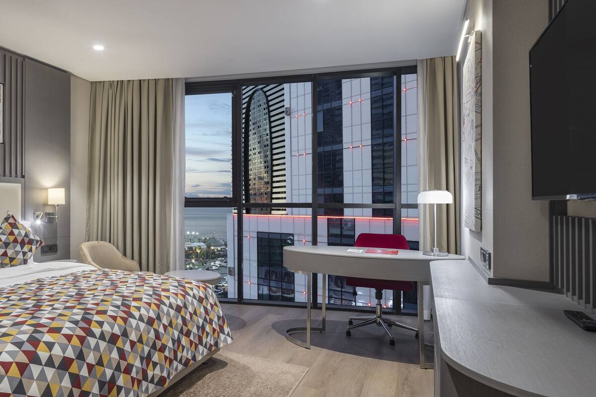 Une chambre d’hôtel moderne dotée d’un grand lit, d’un bureau et offrant une vue sur le paysage urbain grâce à de grandes fenêtres du sol au plafond.
