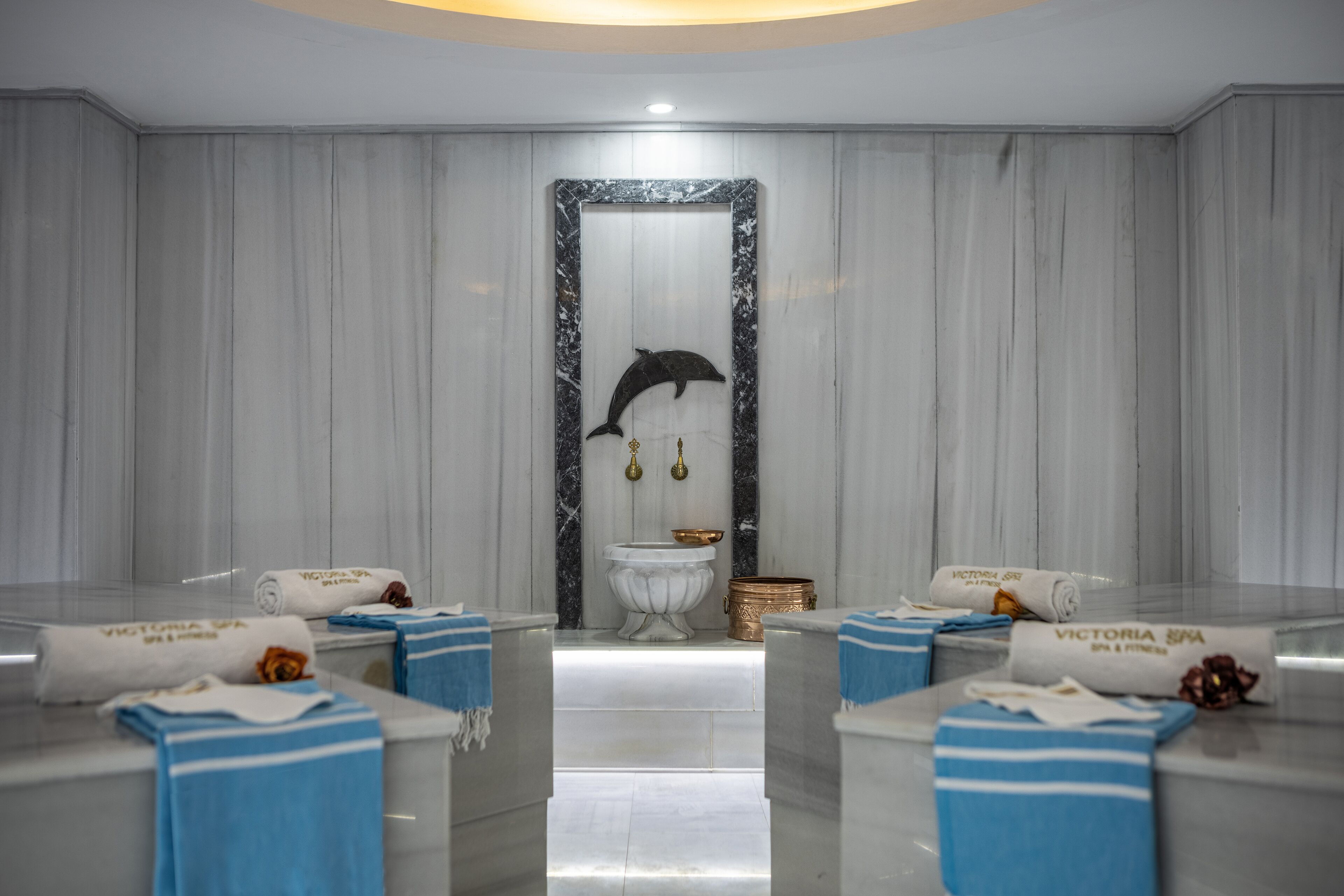 Ein Spa-Raum mit Marmorwand, einer Delphin-Skulptur und Handtüchern mit dem Schriftzug 'Victoria Spa'.