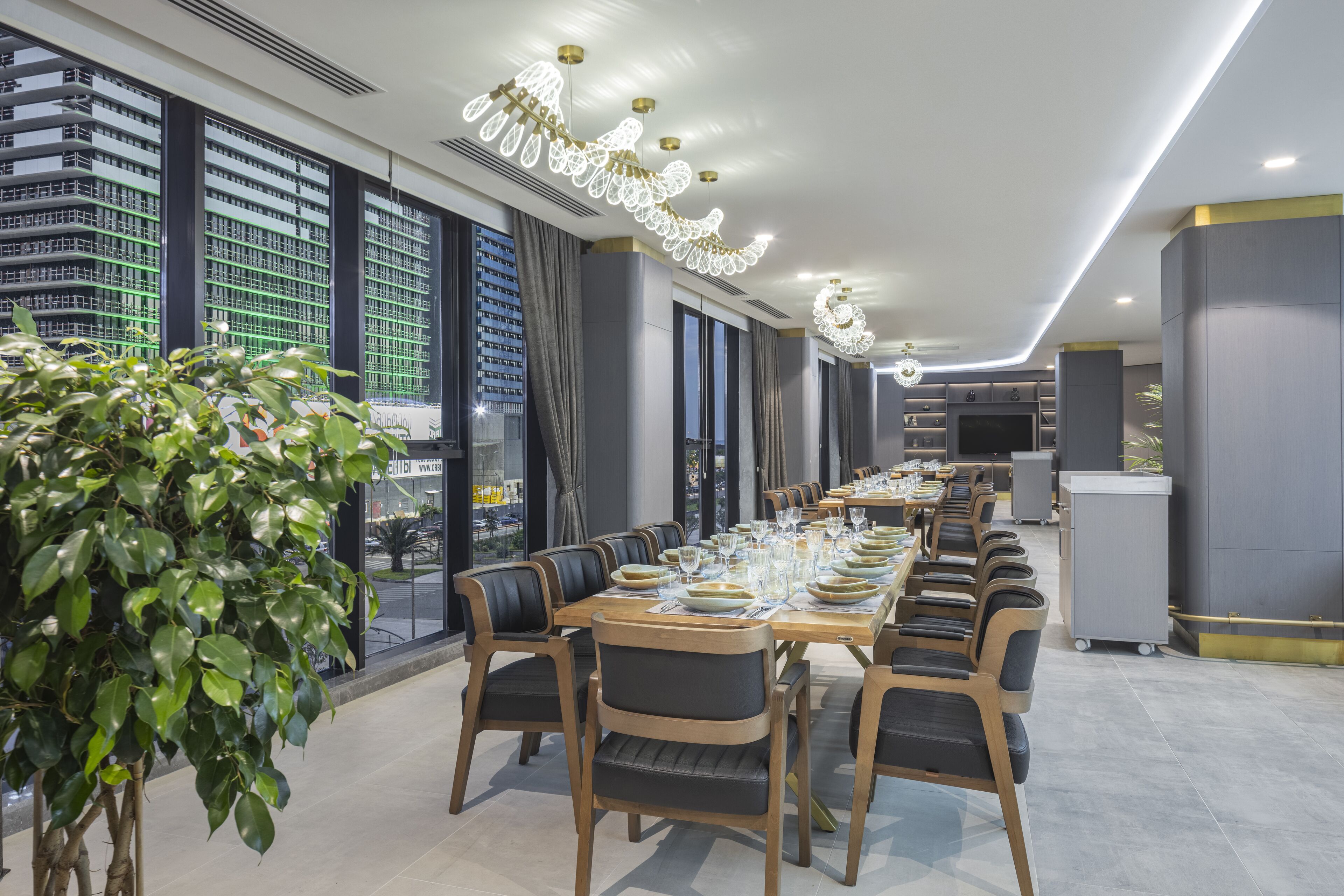 Un comedor moderno con una mesa larga dispuesta para una comida, rodeada de sillas y grandes ventanales con vistas a un paisaje urbano.