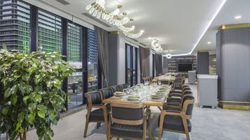 Un comedor moderno con una mesa larga dispuesta para una comida, rodeada de sillas y grandes ventanales con vistas a un paisaje urbano.