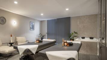 A modern spa room with a massage table, two chairs, a wall clock, a framed artwork, and a glass-enclosed shower area.