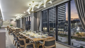 A modern dining area with a long table set for a meal, large windows offering a view of the cityscape, and contemporary lighting fixtures. - Ramada Plaza By Wyndham Batumi (Batumi)