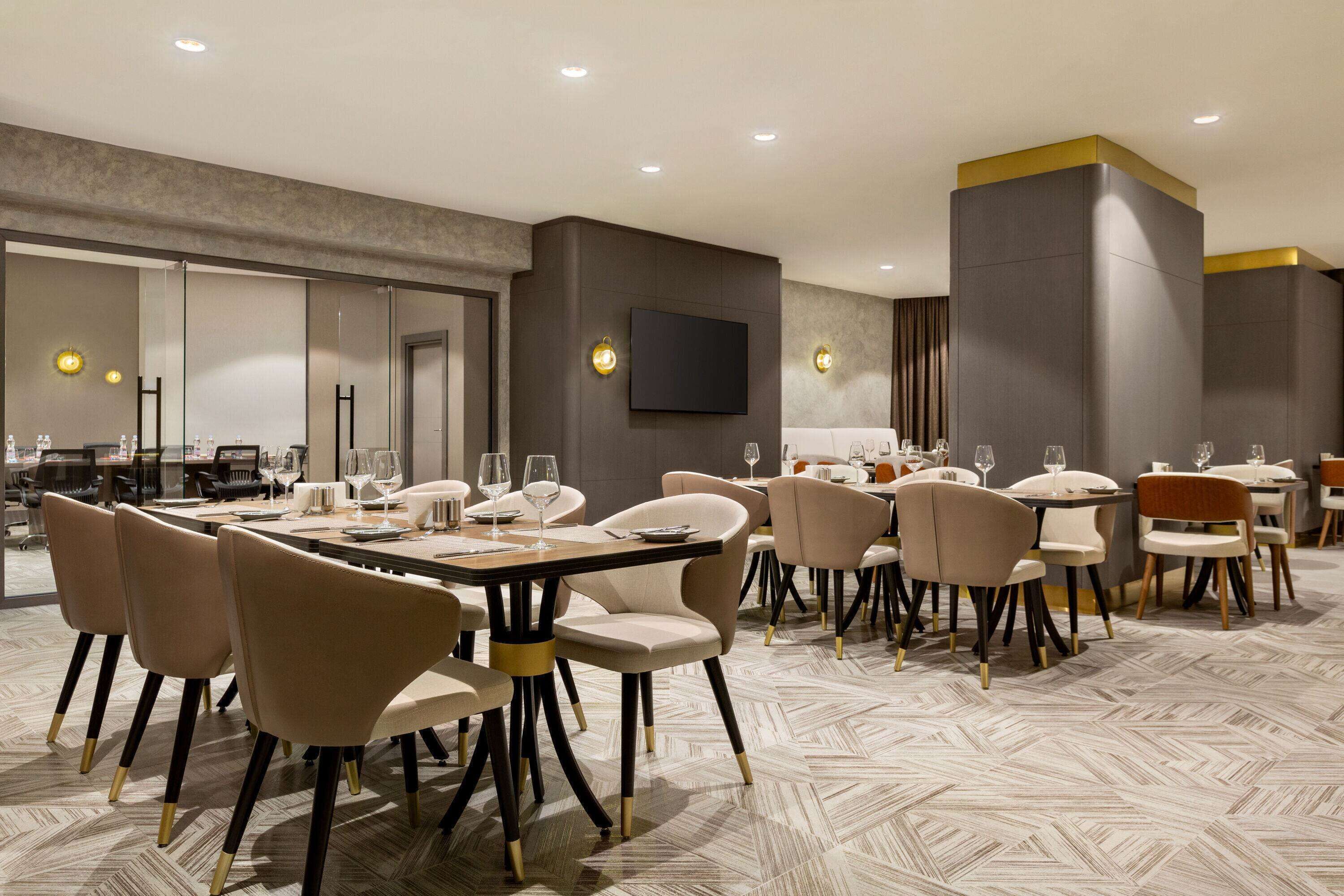 A modern dining area with multiple tables and chairs, a flat-screen TV mounted on the wall, and a patterned floor.