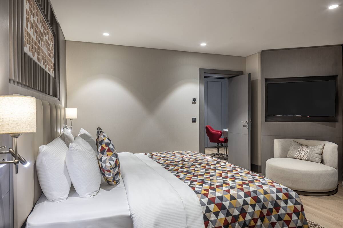 Une chambre d’hôtel moderne avec un grand lit, un téléviseur fixé au mur, un fauteuil rouge et un canapé.