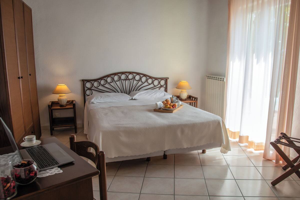 A neatly arranged hotel room with a bed, two bedside tables with lamps, a laptop, and a small table with a fruit bowl.