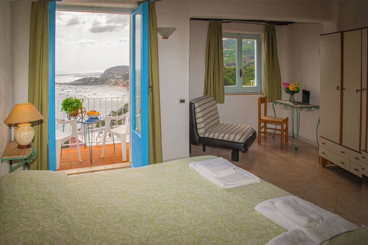 A bedroom with a bed, a chair, a table with a laptop, and a view of the beach.