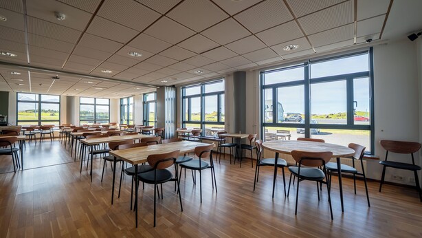 Een ruime binnenzone met meerdere tafels en stoelen, grote ramen met uitzicht op de omgeving, en een hoog plafond met rasterpatronen.