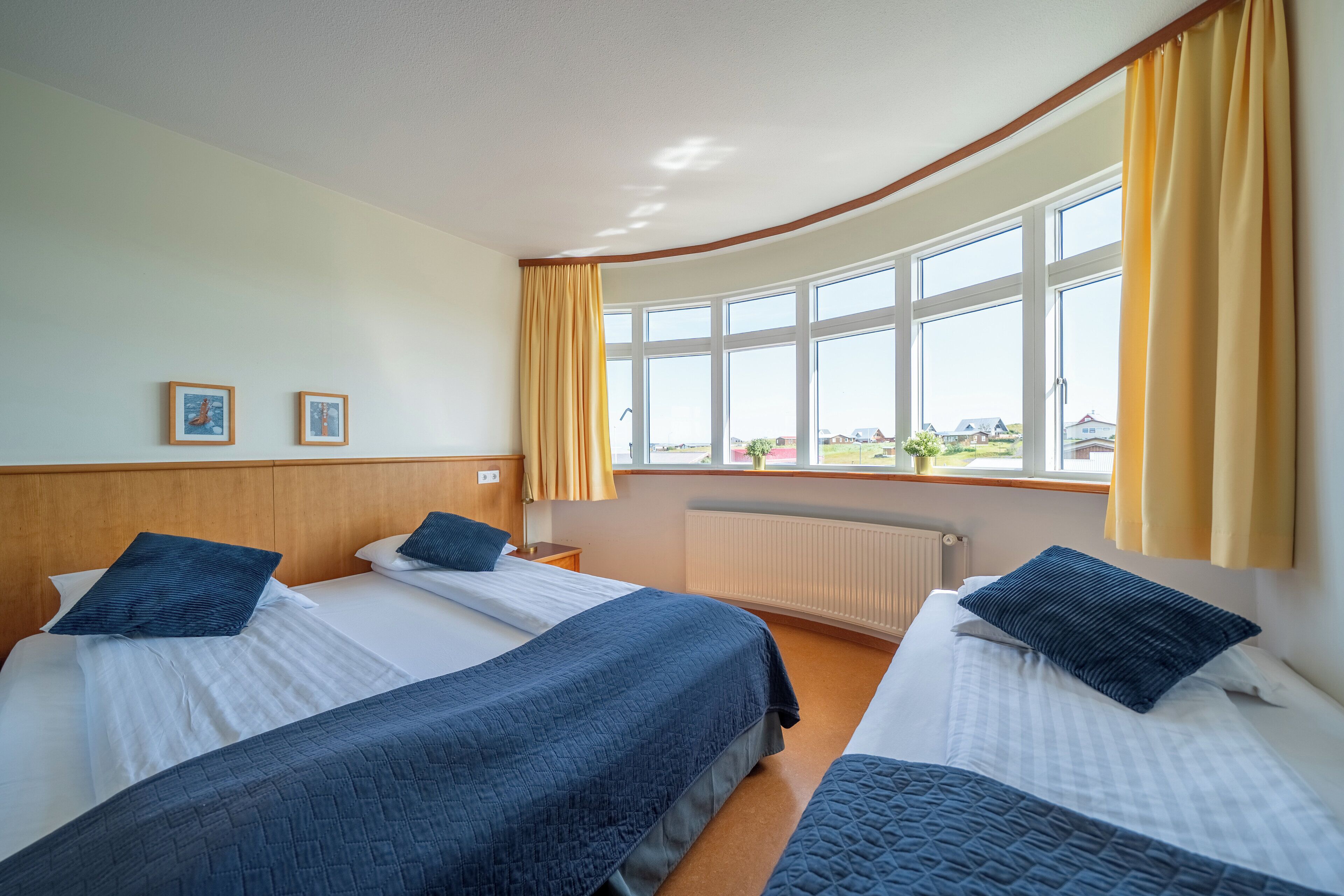 A hotel room with two beds, a large window with curtains, a wooden headboard, and two framed pictures on the wall.