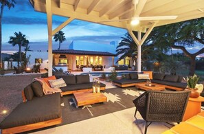 Uma área de estar coberta ao ar livre com sofás e uma mesa de centro, piscina e um edifício com telhado de telhas ao fundo.
