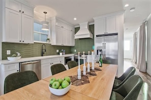 A modern kitchen with white cabinets, a stainless steel refrigerator, and a dining table with a bowl of green apples. - Spacious Mid-Century Oasis | Heated Pool (New Orleans)
