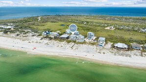 An aerial view of a coastal property with multiple beachfront buildings, a clear view of the ocean, and a dense forested area in the background.