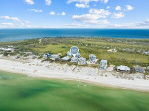 An aerial view of a coastal property with multiple beachfront buildings, a clear view of the ocean, and a dense forested area in the background.