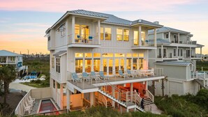 A multi-story beachfront house with a large balcony, outdoor seating, and a swimming pool.