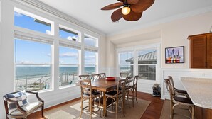 A dining area with a wooden table and chairs, large windows with ocean views, and a ceiling fan.