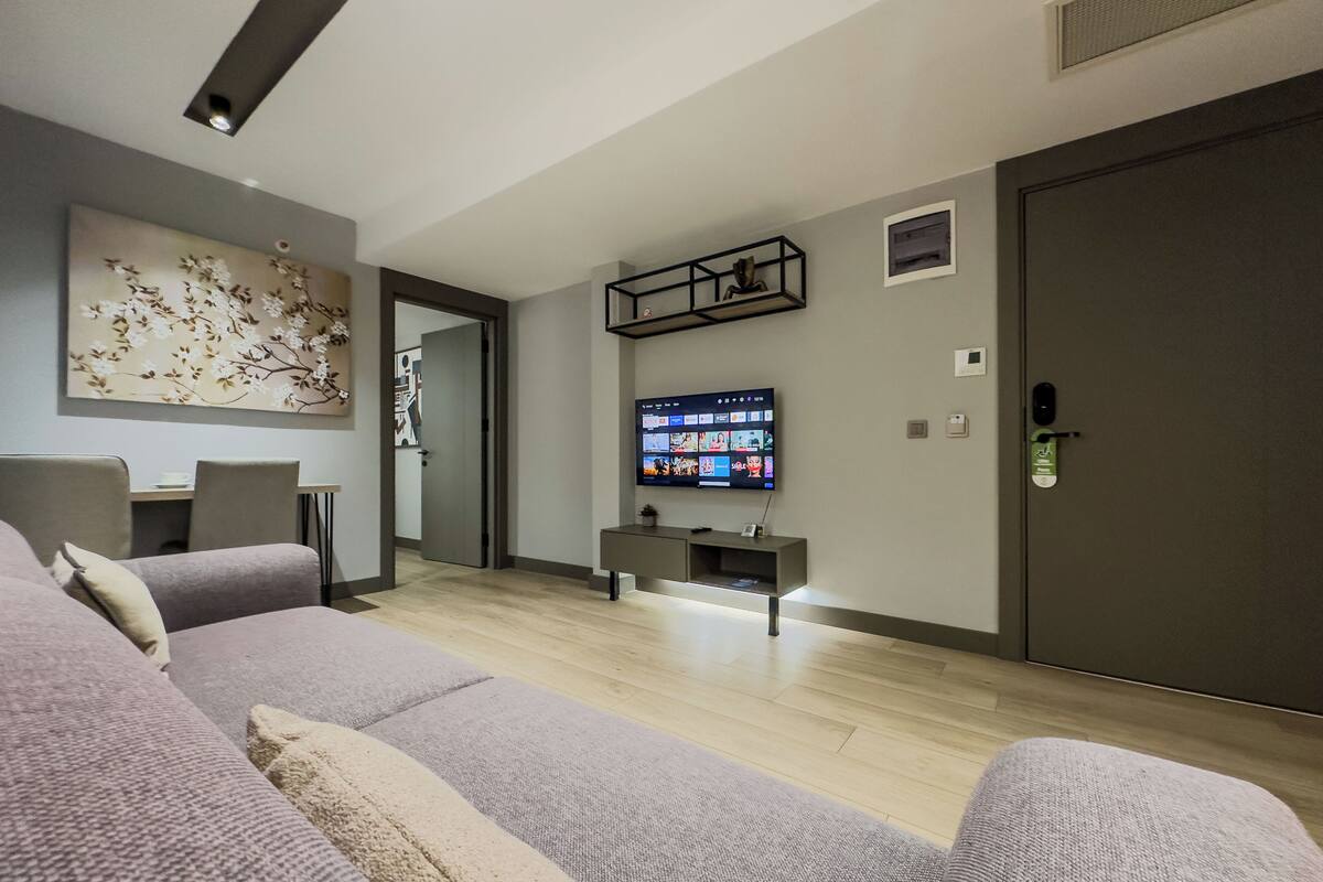 Een moderne woonkamer met een grijze bank, een flatscreen televisie aan de wand en een houten vloer.