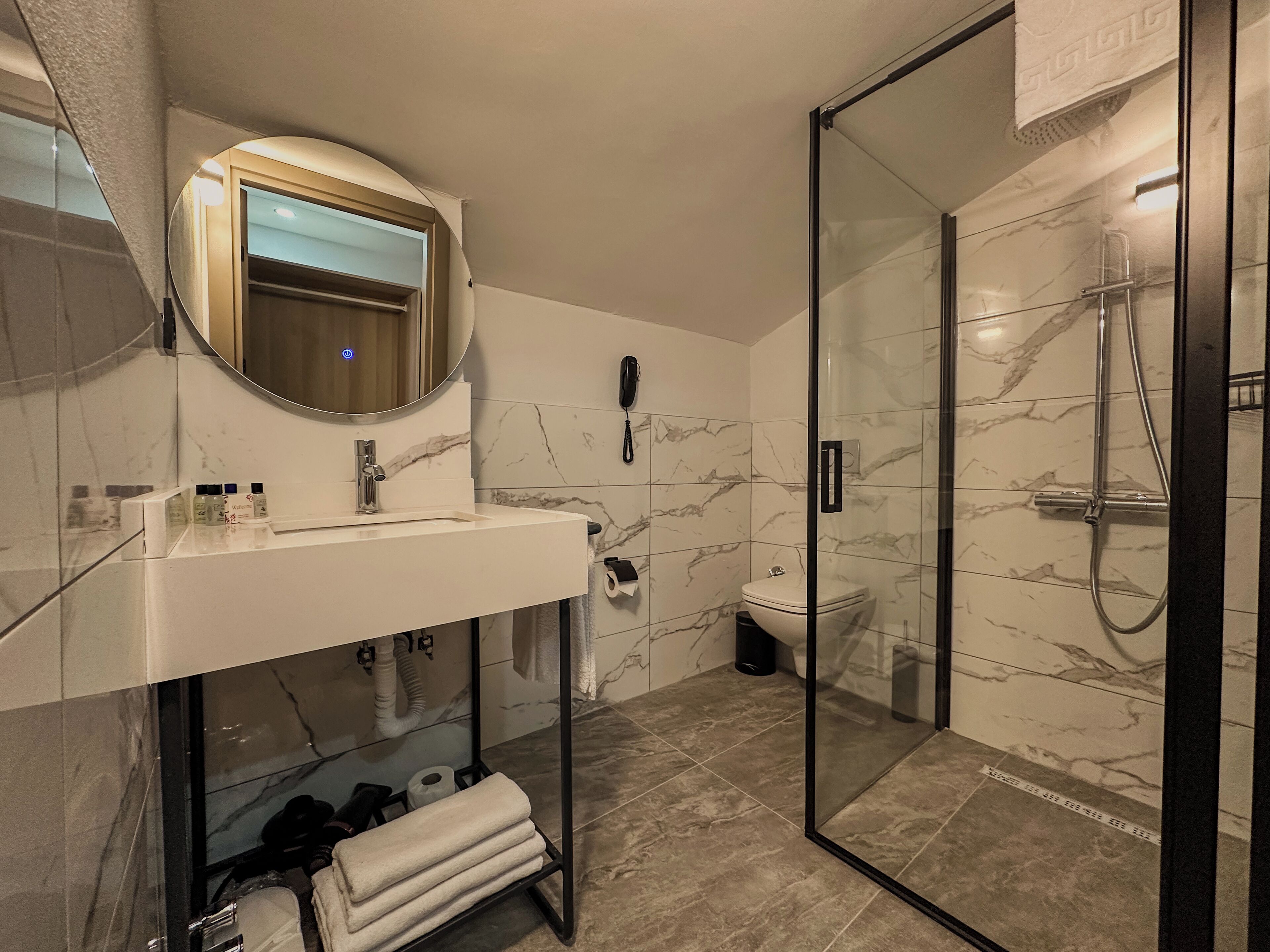Un baño moderno con ducha de vidrio, pared de azulejos de mármol, espejo redondo y lavamanos blanco con artículos de aseo.