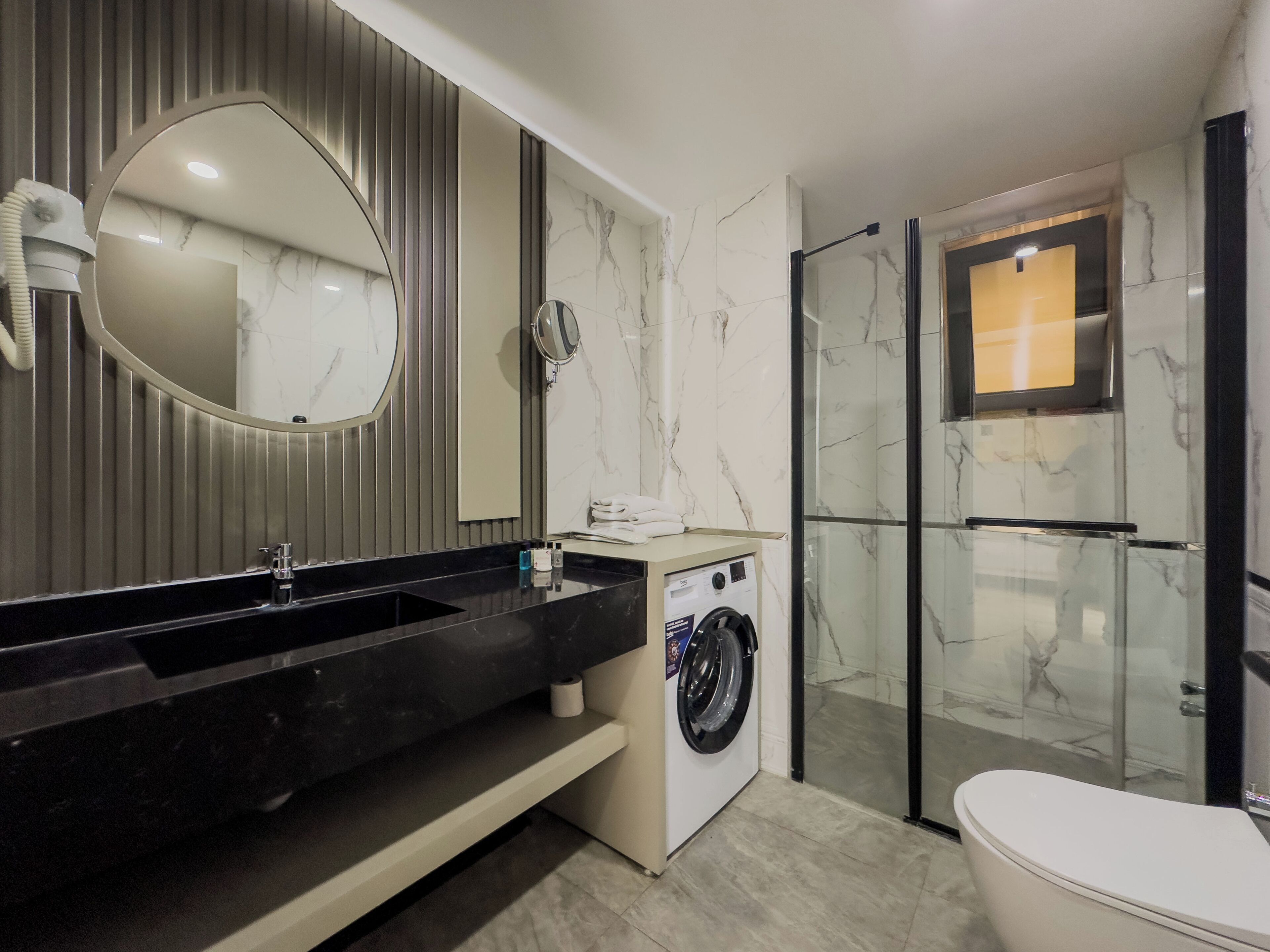 A modern bathroom with a black sink, a built-in washing machine, a glass shower enclosure, and a large mirror.