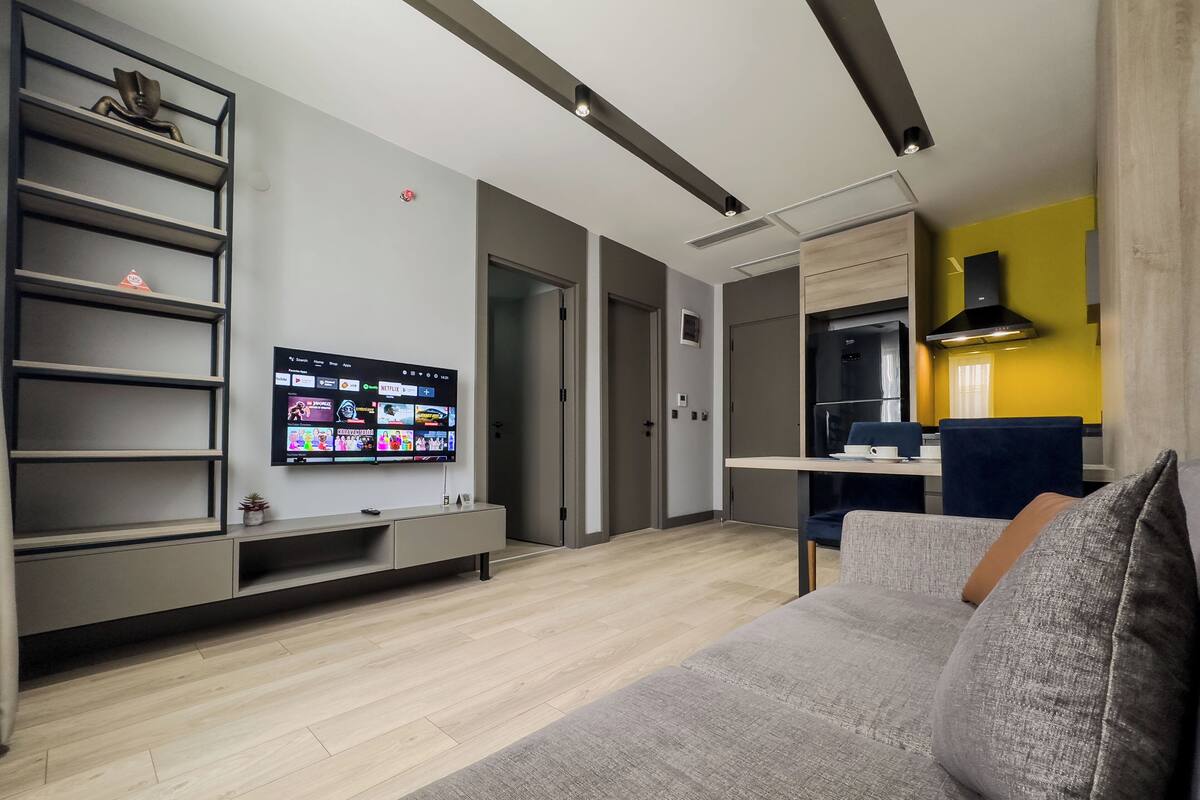 Een moderne woonkamer met een flatscreen televisie aan de muur, een grijze bank en een houten vloer.