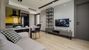 Een moderne woonkamer met een bank, een eettafel met stoelen, een aan de muur bevestigde televisie en een keukenhoek met een gele wand en zwarte kastjes.
