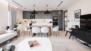 Ein modernes Wohnzimmer mit Sofa, Couchtisch und Fernseher. Ein Essbereich mit Tisch und Stühlen ist ebenfalls vorhanden. Die Küche ist sichtbar, mit Schränken und einer Arbeitsplatte.