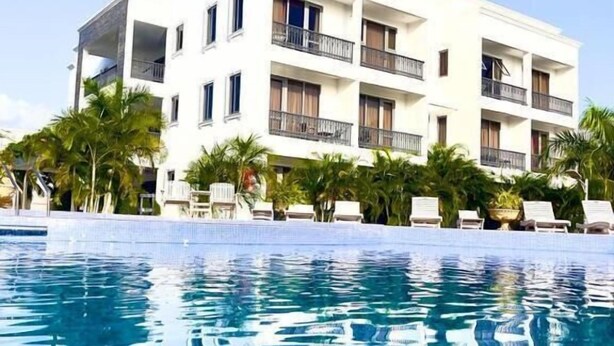 Um hotel branco de vários andares com varandas, uma piscina em primeiro plano e palmeiras ao redor.