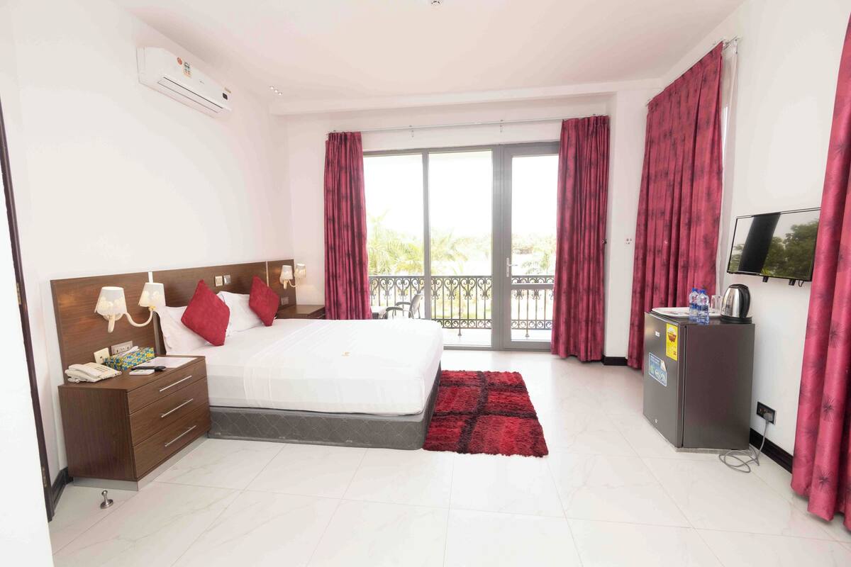 A hotel room with a bed, a nightstand, a desk with a kettle, a TV, and a balcony with red curtains.