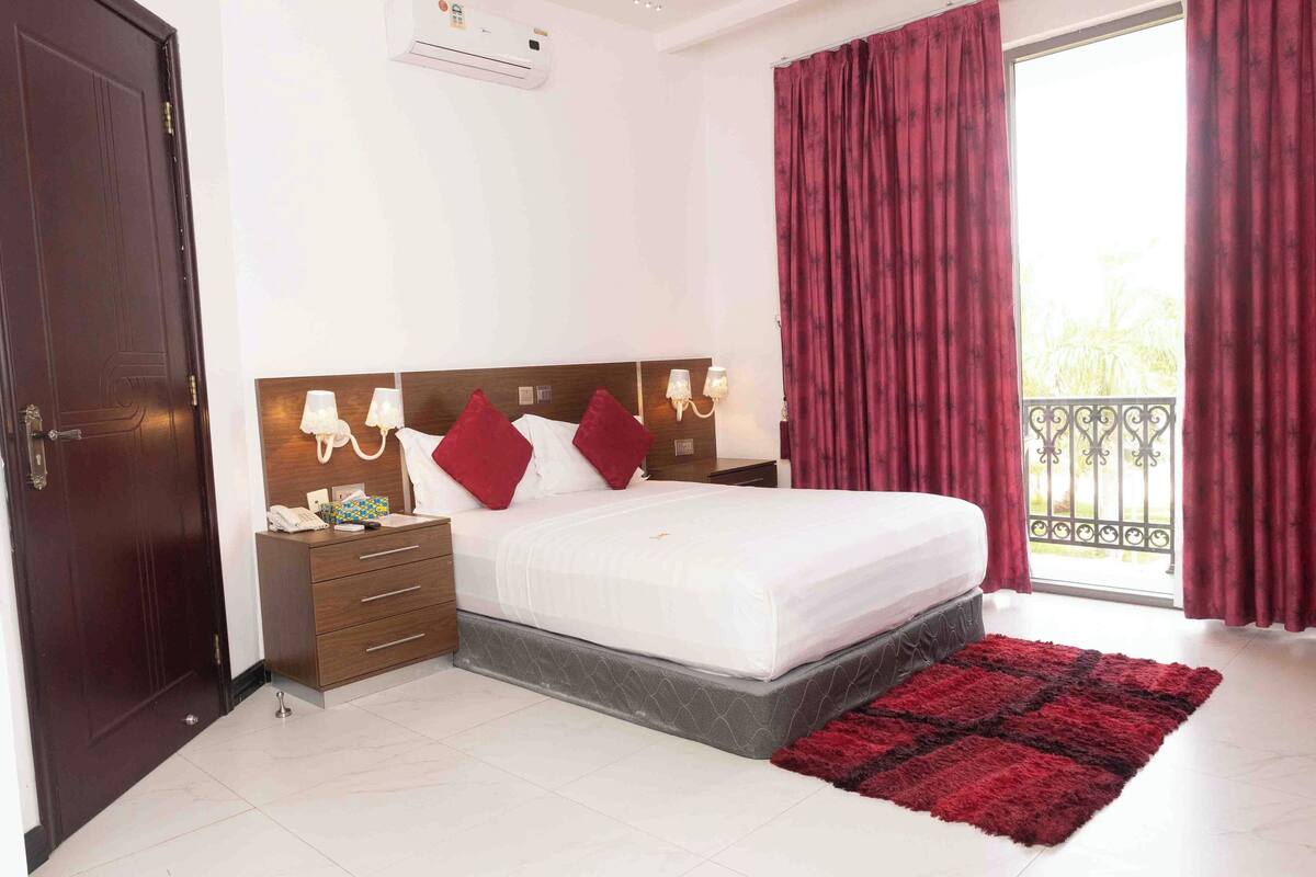 A hotel room with a large bed, a wooden nightstand, a red rug, and a balcony with red curtains.