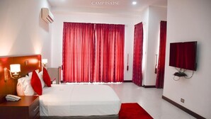 Quarto de hotel com uma cama grande, mesa de cabeceira com abajur, televisão na parede e cortinas vermelhas.