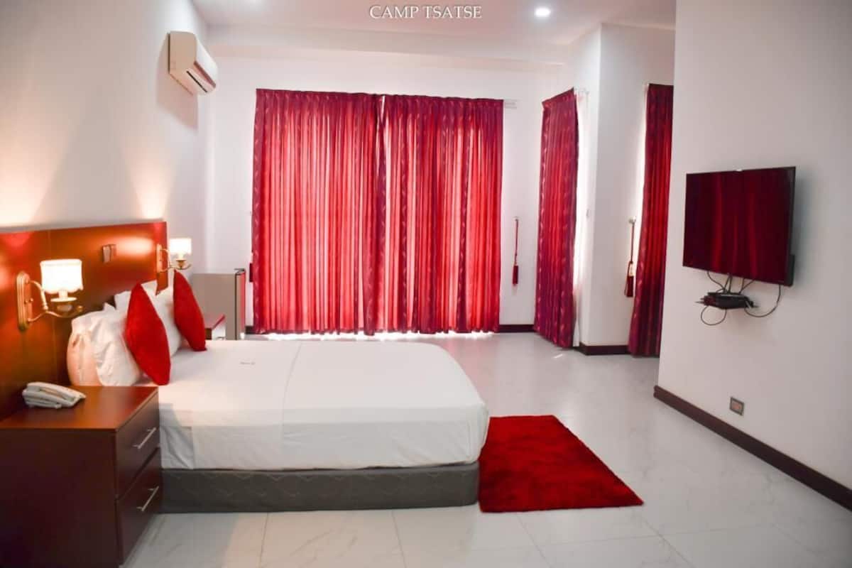 A hotel room with a large bed, a bedside table with a lamp, a TV mounted on the wall, and red curtains.