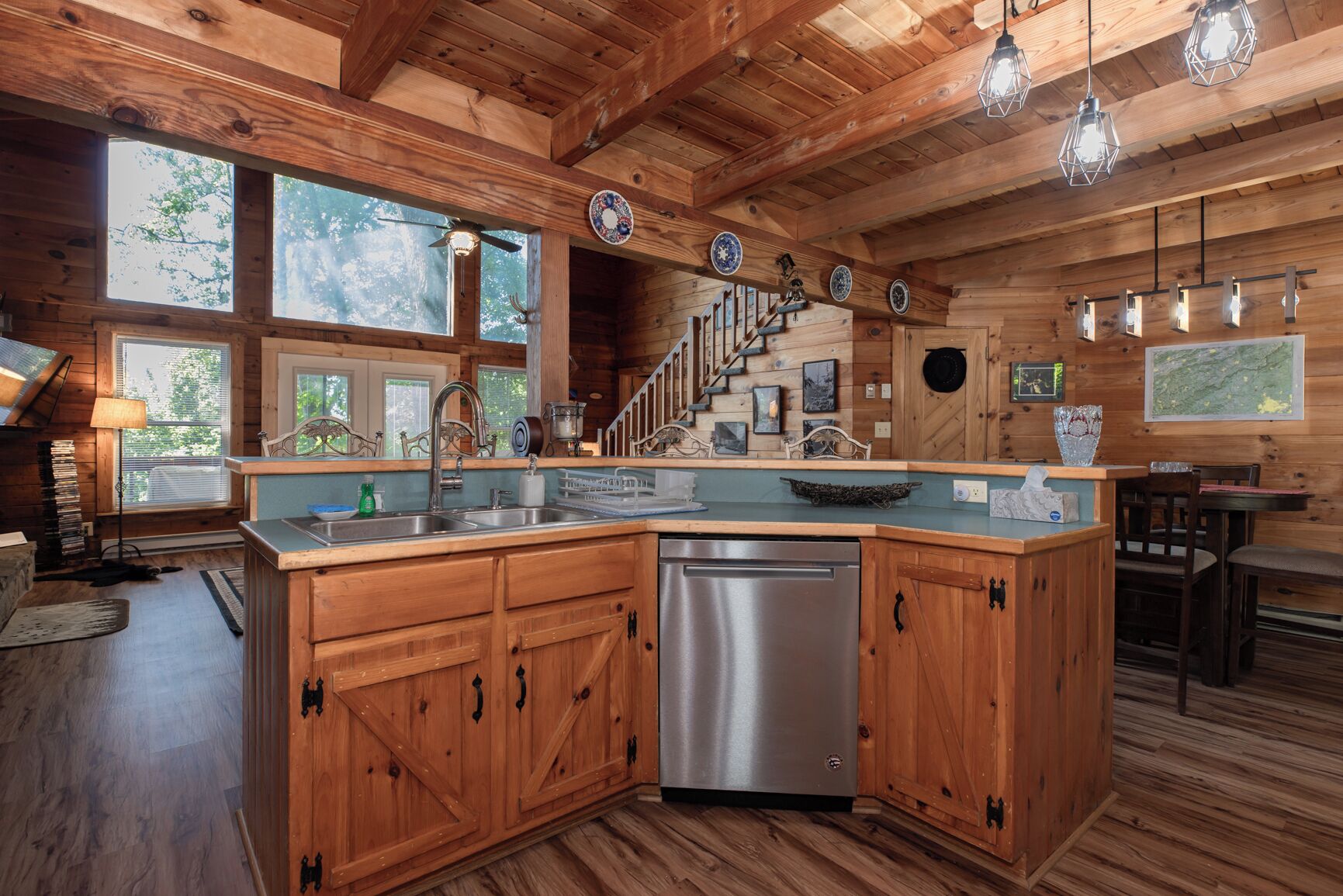 A wooden cabin with a kitchen featuring a stainless steel dishwasher, wooden cabinets, and a large window overlooking the outdoors.