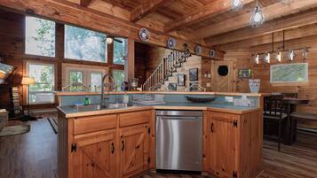 A wooden cabin with a kitchen featuring a stainless steel dishwasher, wooden cabinets, and a large window overlooking the outdoors.