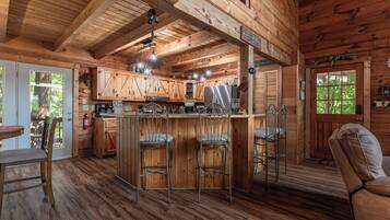 A rustic cabin interior with a wooden bar, bar stools, a dining table, and a cozy seating area.