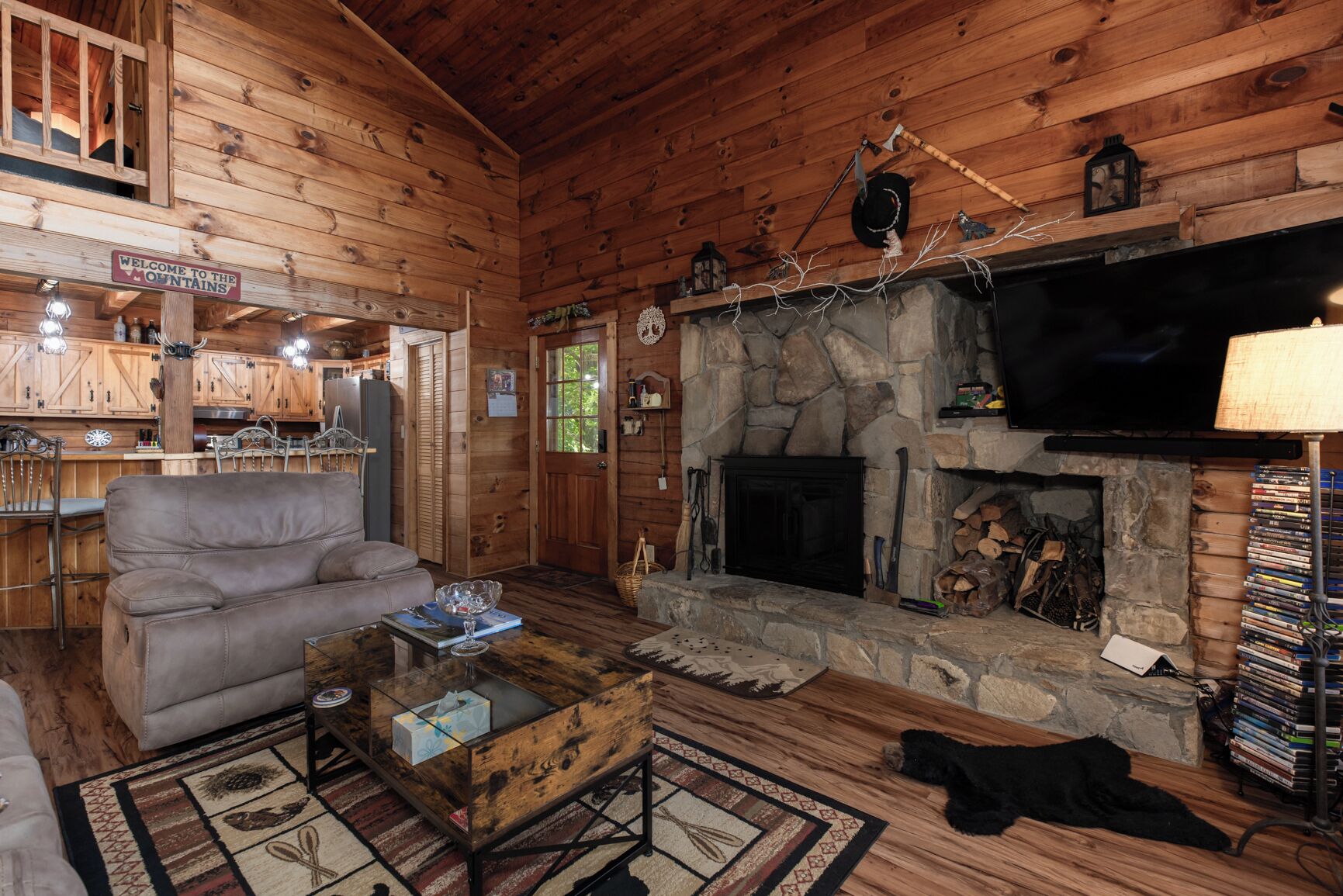 A cozy log cabin interior with a stone fireplace, a television mounted above it, and a bar area with wooden cabinets and a sign reading "Welcome to the Mountains".
