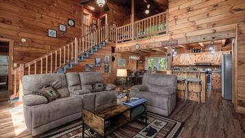 A cozy cabin interior with a wooden staircase, a living area with a sofa and armchairs, a dining area, and a kitchen.