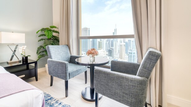 A hotel room with a large window, two grey armchairs, a small round table with a vase of flowers, a black nightstand with a lamp, and a potted plant.