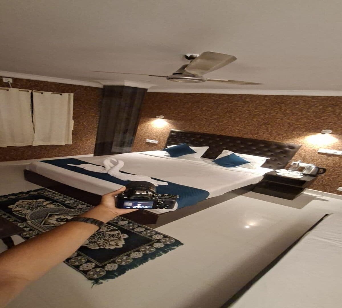 A hotel room with a large bed, a ceiling fan, a nightstand, and a patterned rug.
