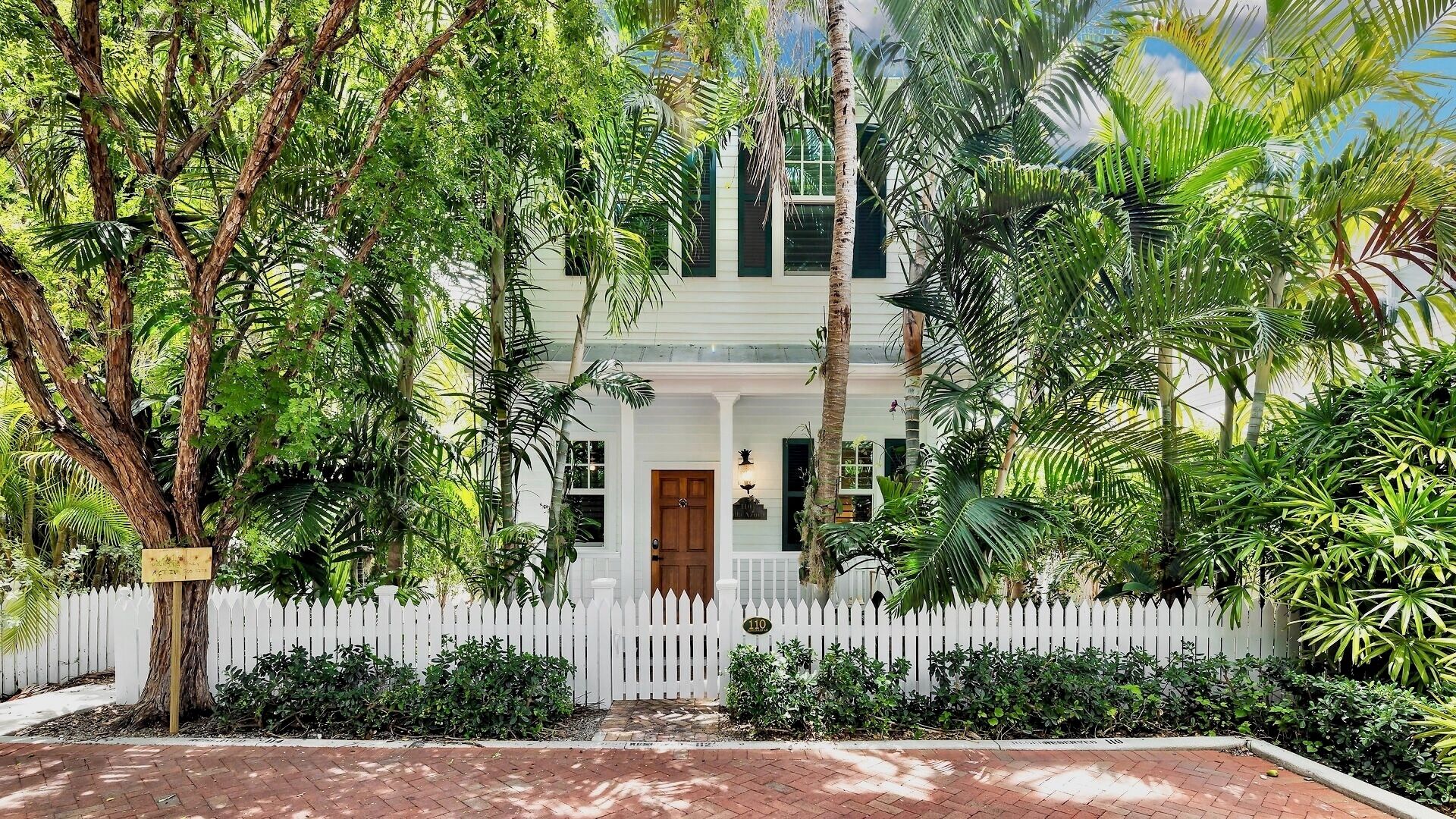 A two-story house with a white picket fence, surrounded by lush greenery and palm trees.