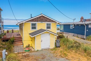 Une petite maison jaune, avec une porte blanche et un petit porche, située près d’une plage, avec une maison bleue en arrière-plan.