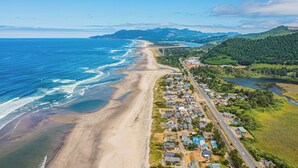 Vista aérea de una zona costera con playa, una carretera bordeada de casas y una colina boscosa al fondo.