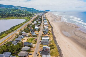 Vue aérienne d’une zone résidentielle côtière, avec des maisons le long de la plage, une route parallèle au rivage et une étendue d’eau sur la gauche.