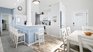 A modern kitchen with white cabinetry, a central island, and a dining area with white chairs. - Sunset Overlook, Spacious bay side top-floor condo with amazing views  (Ocean City)